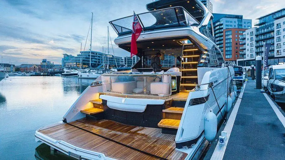 Luxury yacht docked at a marina, featuring wooden decking, lounge area, and a red flag; city buildings in the background.